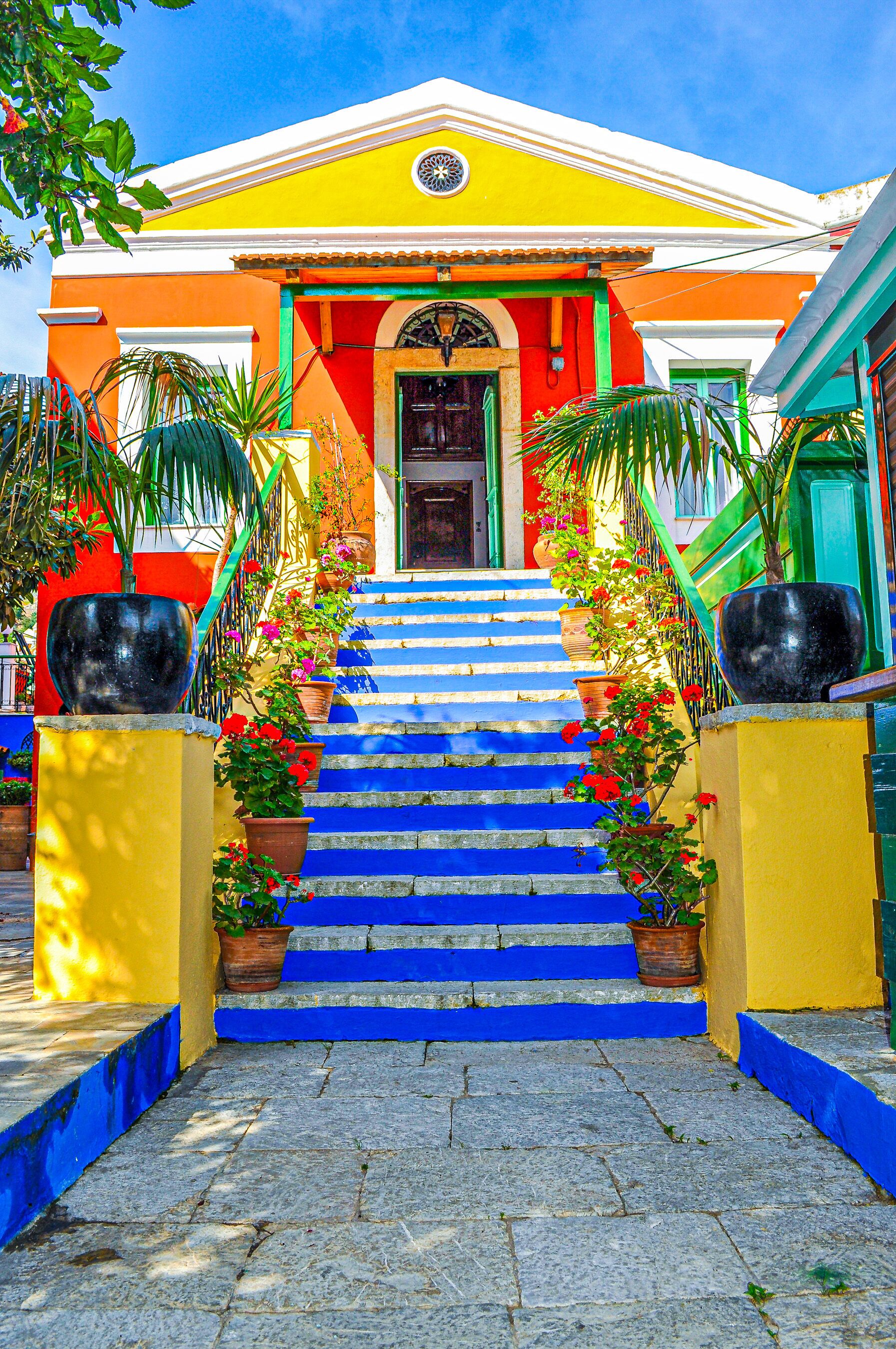 A brightly colored building with a staircase leading to the entrance, surrounded by potted plants and a clear blue sky.