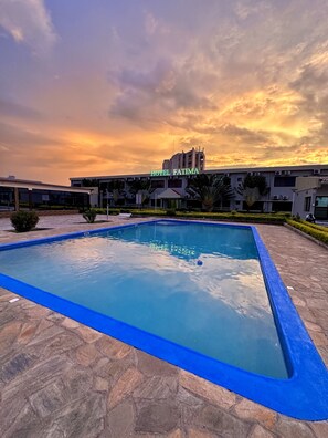 Una piscina con borde azul, rodeada de baldosas de piedra, y un edificio de hotel al fondo.