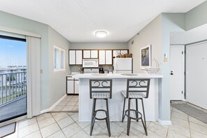 A kitchen with a white island, two bar stools, a microwave, and a refrigerator. - Plantation Dunes Condos (Gulf Shores)