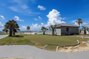 A golf course with a putting green, palm trees, and a building with a gray roof. - Plantation Dunes Condos (Gulf Shores)