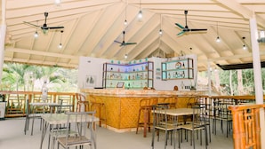 A restaurant with a high ceiling, multiple ceiling fans, and a bar area with shelves of bottles. Tables and chairs are arranged for dining. - Denika's Suites (Puerto Princesa)