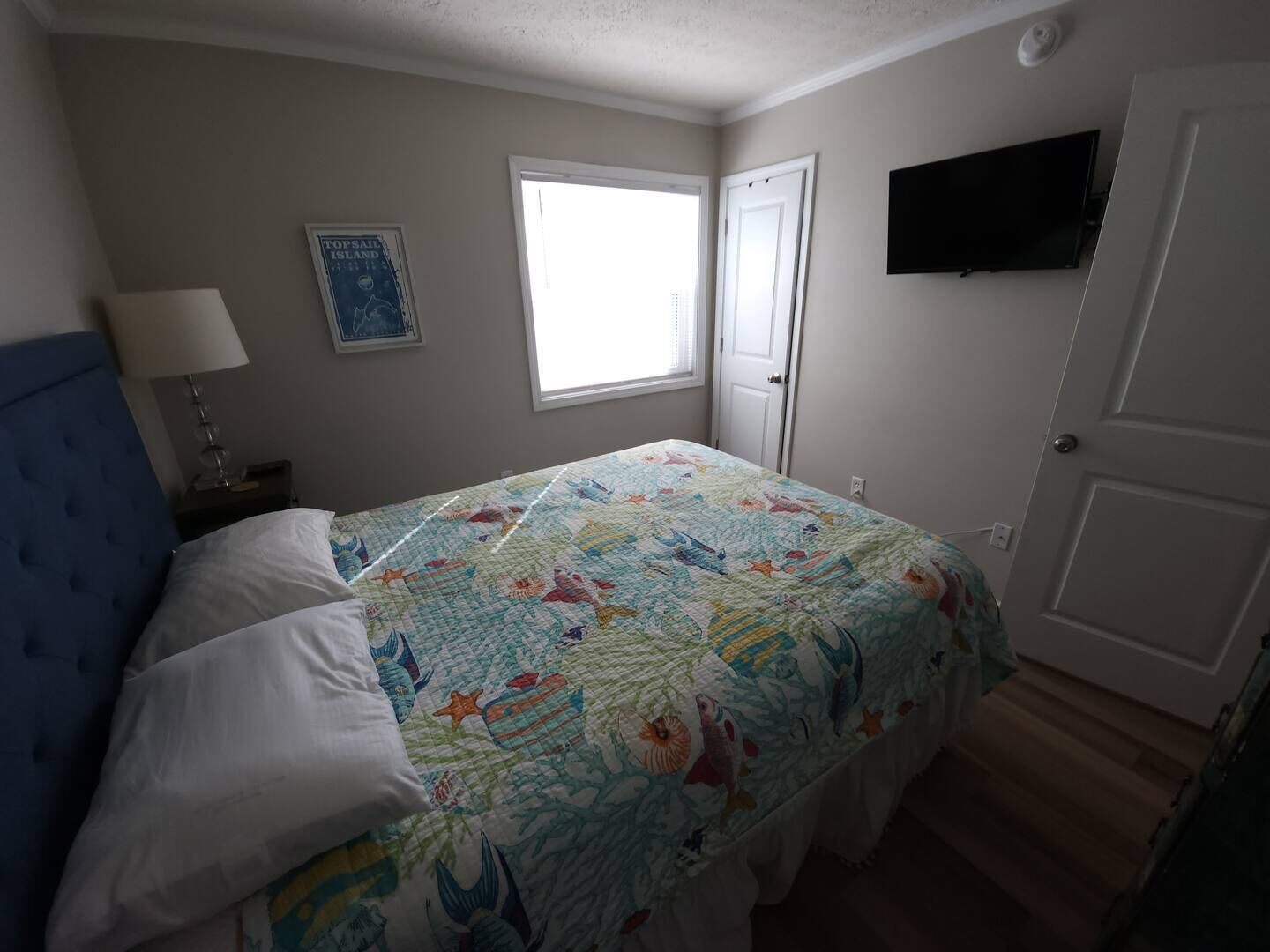 Een slaapkamer met een bed, een raam, een deur, een aan de muur bevestigde televisie en een poster.
