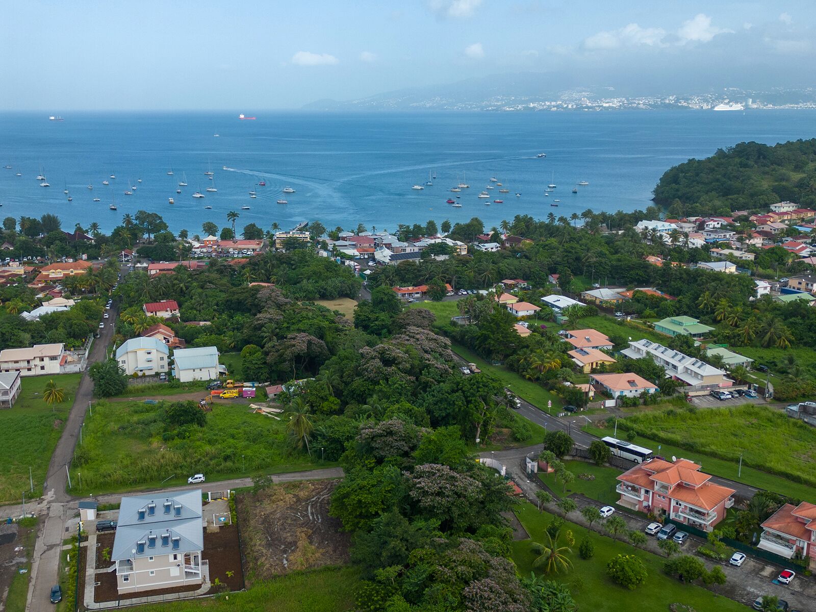 Vue aérienne d’une ville côtière avec des immeubles résidentiels, une marina remplie de bateaux et, au loin, un paysage vallonné.