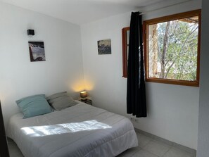 Ein Schlafzimmer mit Bett, Nachttisch, Wandlampe, zwei Wandbildern und einem Fenster mit Vorhängen.
