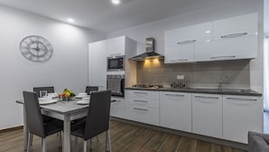 Une cuisine moderne avec des meubles blancs, un four encastré, un four à micro-ondes et une cuisinière à gaz. On y trouve une table à manger avec des chaises et un panier de fruits.