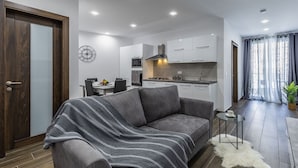 Un salon moderne avec un canapé gris, une table à manger avec des chaises, un coin cuisine avec des meubles blancs et une grande fenêtre avec des rideaux.