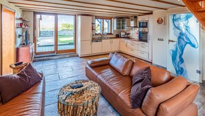 A spacious living room with a brown leather sofa, a glass coffee table with wood logs, and a view of the kitchen and outdoor area through large sliding glass doors. - Cozy home in Lübeck with house sea view (Lübeck)