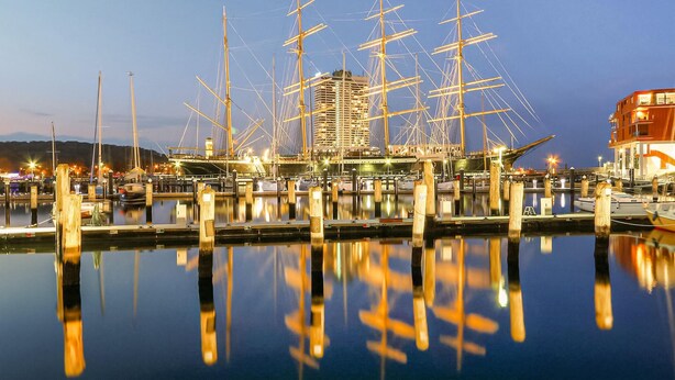 A marina with multiple tall ships docked, a modern building in the background, and calm water reflecting the ships. - Cozy home in Lübeck with house sea view (Lübeck)
