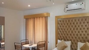 Een hotelkamer met een gestoffeerd hoofdeinde, een bed met kussens, een ronde tafel met stoelen, een raam met zonwering en een airconditioner aan de muur.