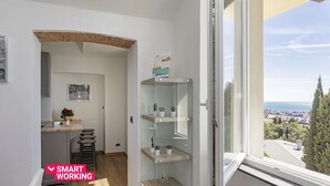 Une petite pièce lumineuse donnant sur un paysage urbain, avec un bureau en bois et une chaise, ainsi qu’une étagère où sont disposées des plantes.