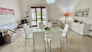 Amplia sala de estar con una mesa de comedor de vidrio, sillas blancas, un sofá y un televisor.