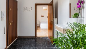 Um corredor com porta de madeira, parede branca com luminária, e banheiro com pia e espelho brancos.