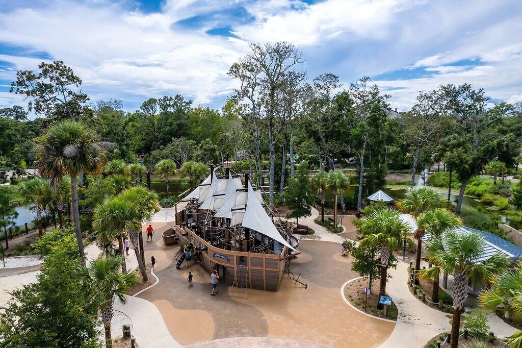 Una amplia estructura al aire libre con techo curvo, rodeada de palmeras y otra vegetación.