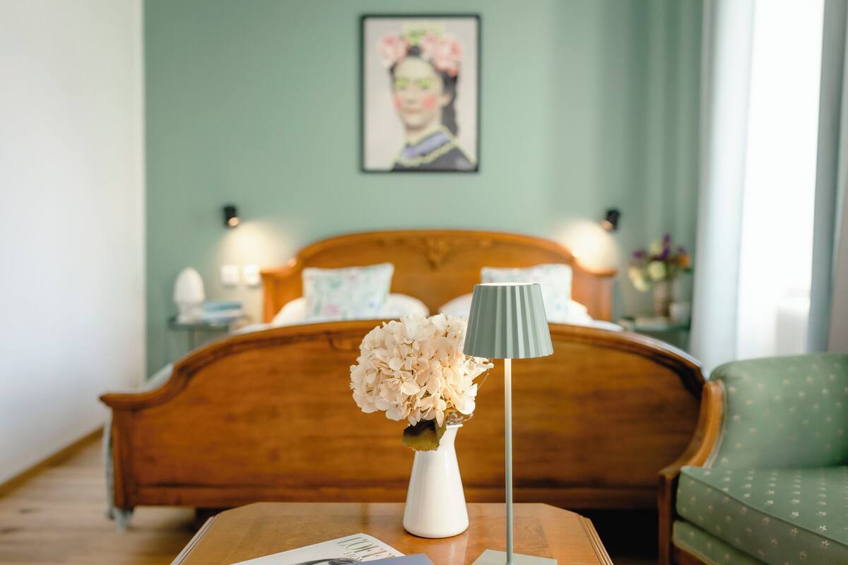 A bedroom with a wooden bed, a white vase with flowers, a lamp, and a green chair.