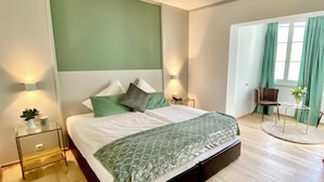 A modern bedroom with a large bed, green accent pillows, a bedside table with a lamp, and a view of a balcony with curtains. - Boutiquehotel Hubertushof (Bad Ischl)