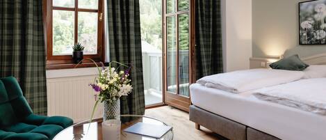 A hotel room with a bed, a round glass table with a laptop, a green armchair, and a window with curtains.