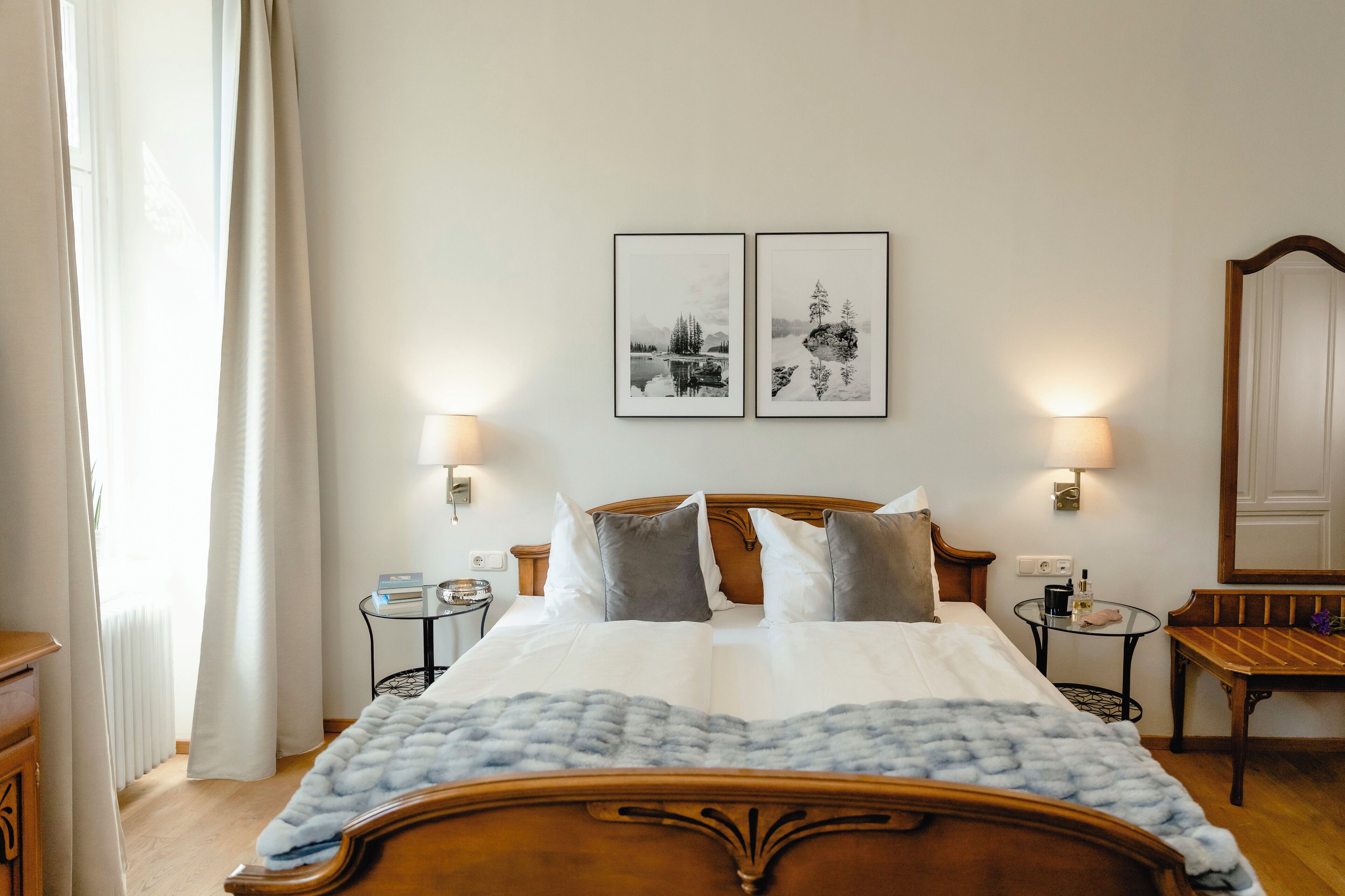 Un letto rifatto con cura, una testiera in legno, due comodini con lampade, un tavolino tondo con lampada e due quadri appesi al muro.