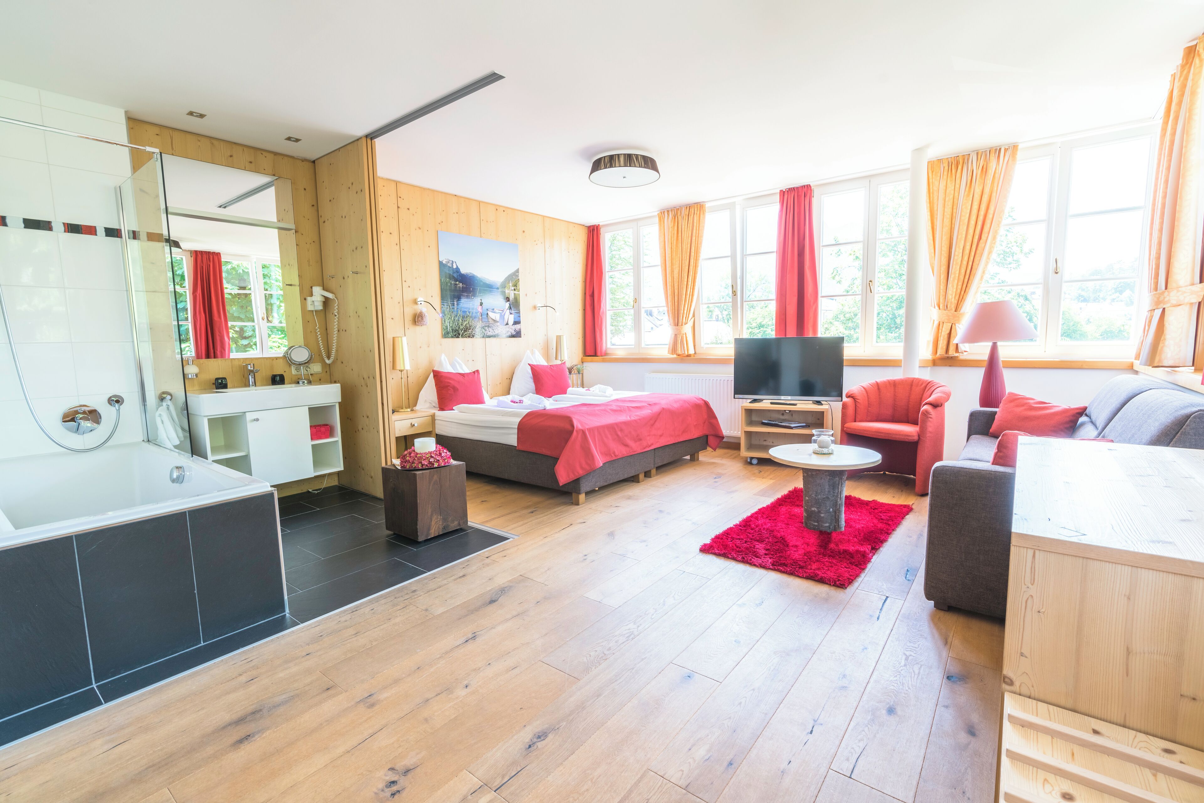 Habitación de hotel moderna con detalles de madera, una cama grande, televisión, escritorio y zona de estar con un tapete rojo.