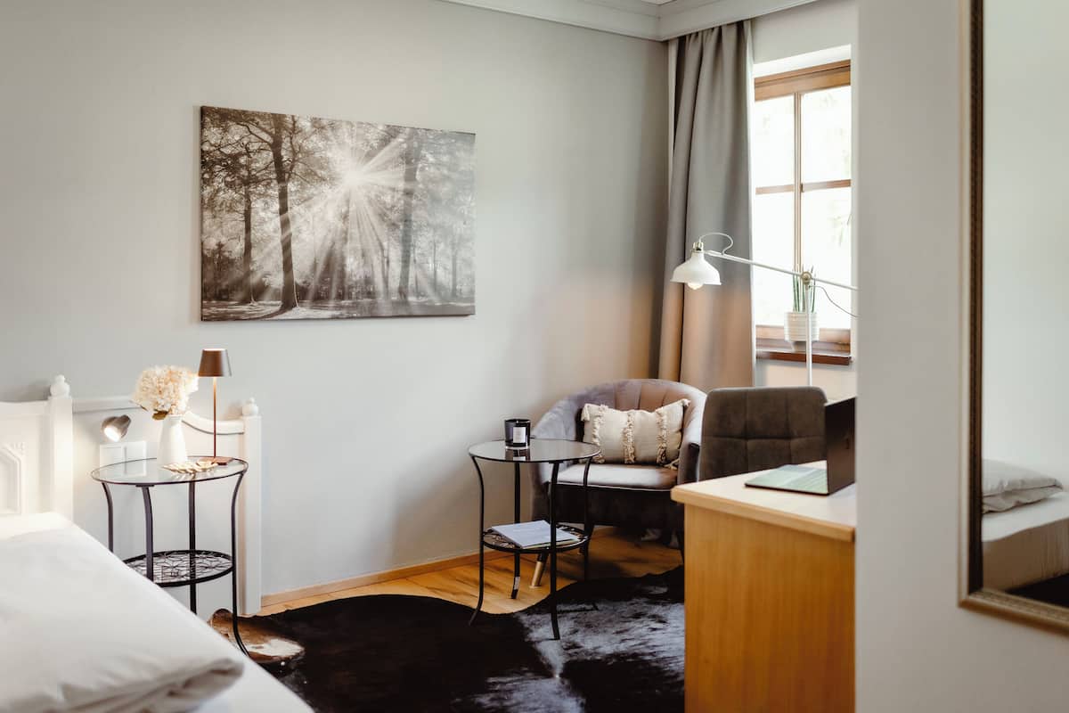 A modern hotel room with a bed, a desk with a laptop, a chair, a small round table with a lamp, and a large forest-themed painting on the wall.