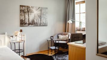 A modern hotel room with a bed, a desk with a laptop, a chair, a small round table with a lamp, and a large forest-themed painting on the wall.