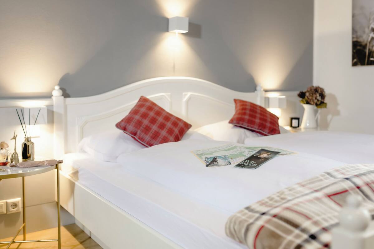A neatly made bed with white linens, two red plaid pillows, and a magazine on the bed. There is a small table with a lamp and a vase of flowers to the left of the bed.