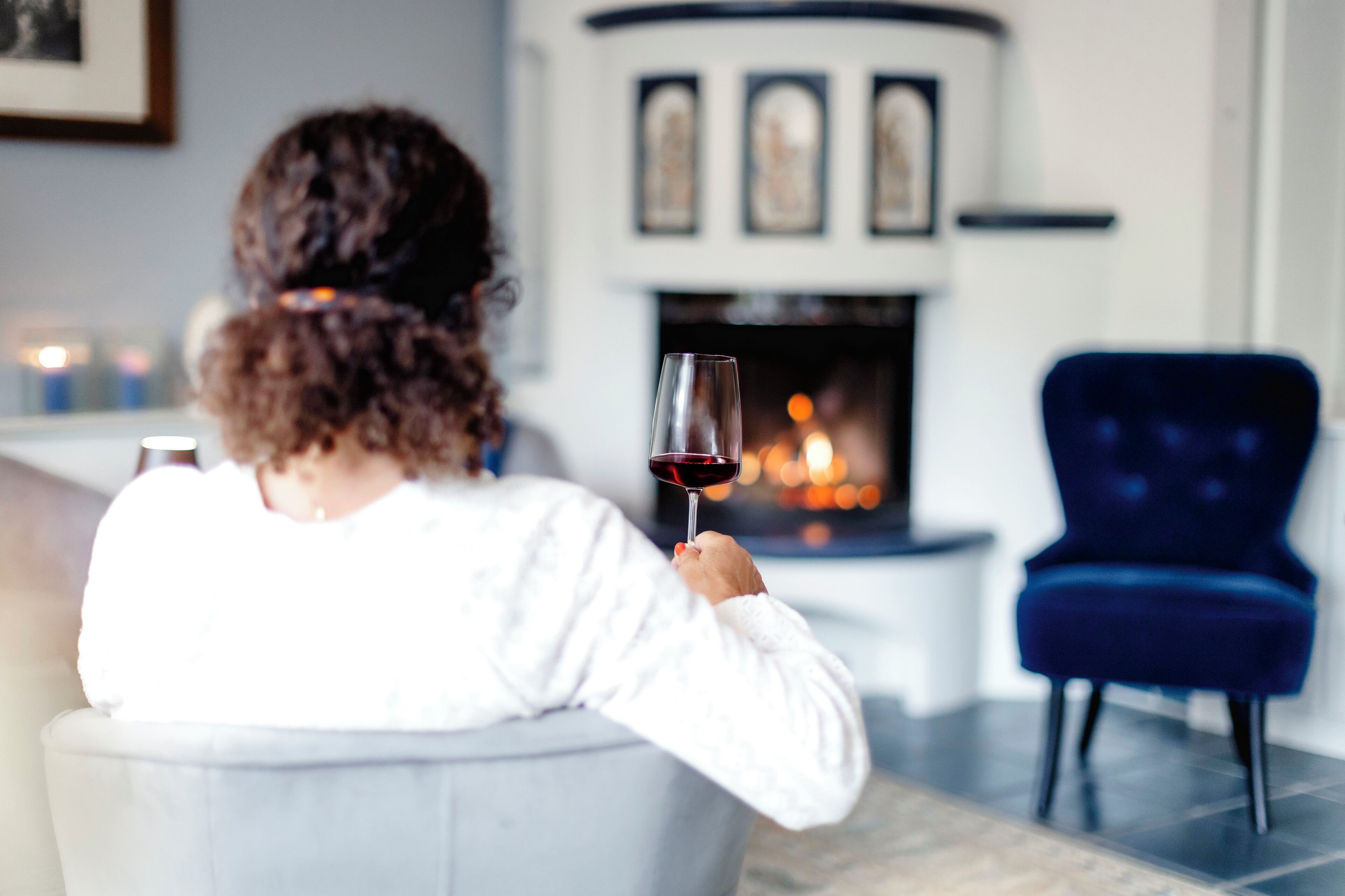 A person seated on a white chair in a room with a fireplace, a blue armchair, and framed pictures on the wall.