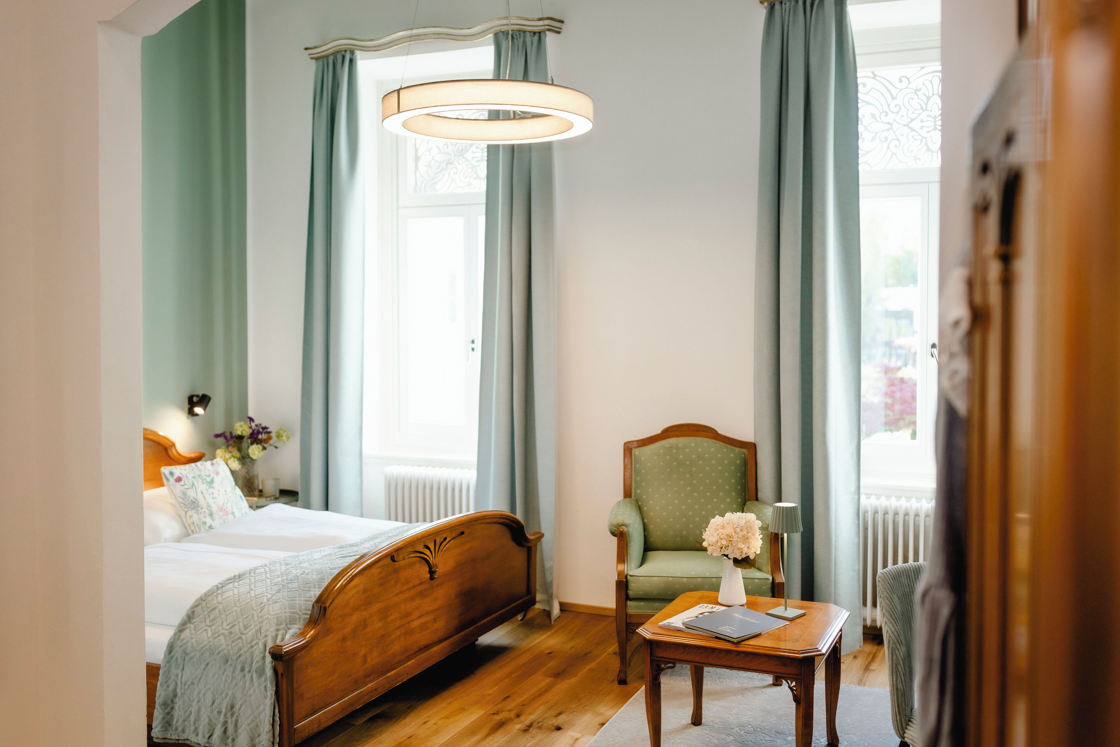Ein Schlafzimmer mit einem Holzbett, einem grünen Sessel, einem kleinen Tisch mit einer Vase und einem Fenster mit Vorhängen.