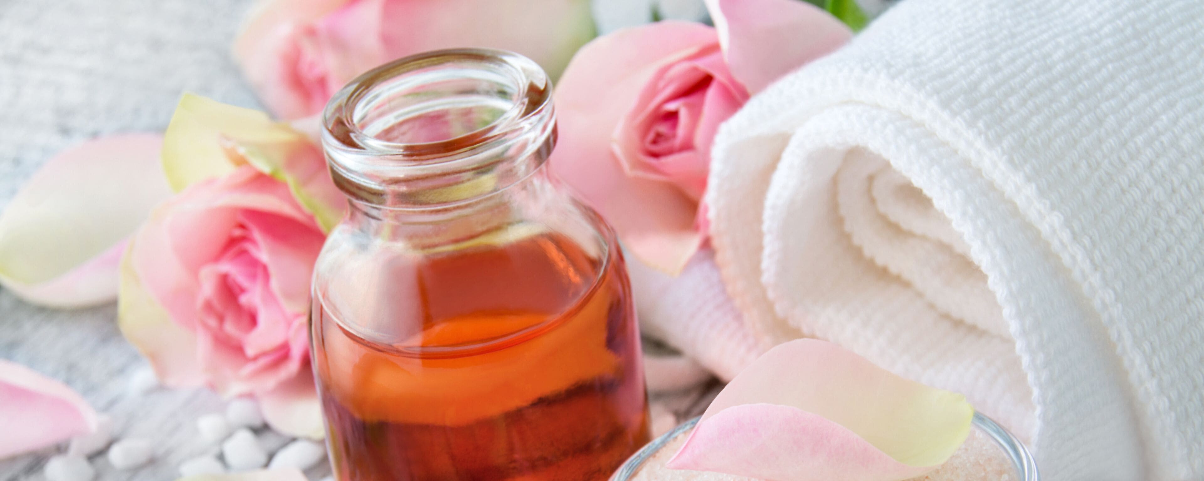 A bottle of amber liquid, pink rose petals, and a rolled white towel.