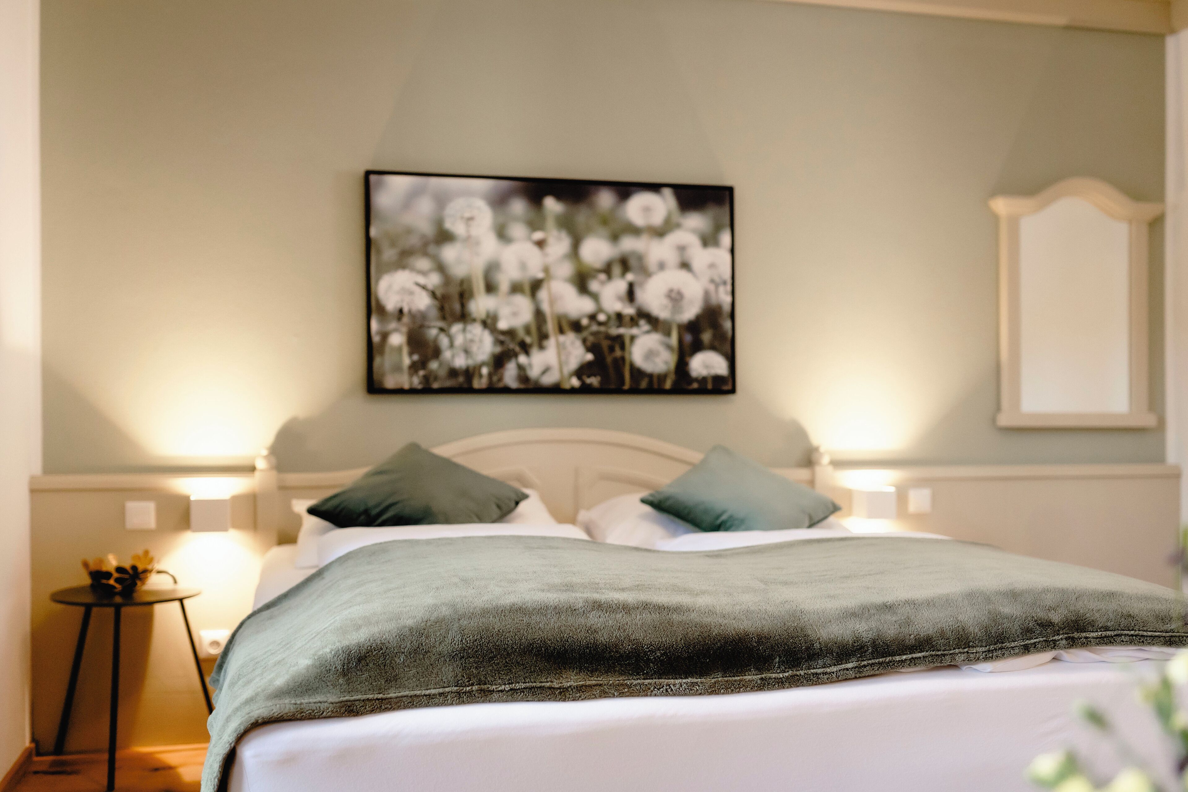 Una cama bien tendida con una manta verde, dos almohadas y un cuadro de flores colgado en la pared.