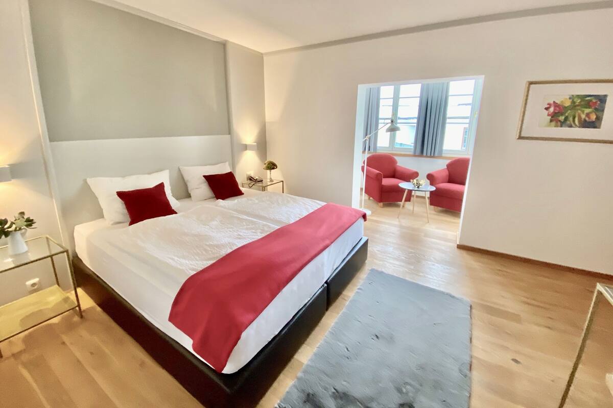 A modern hotel room with a large bed, a bedside table, a nightstand, a desk, a chair, and a window with curtains.