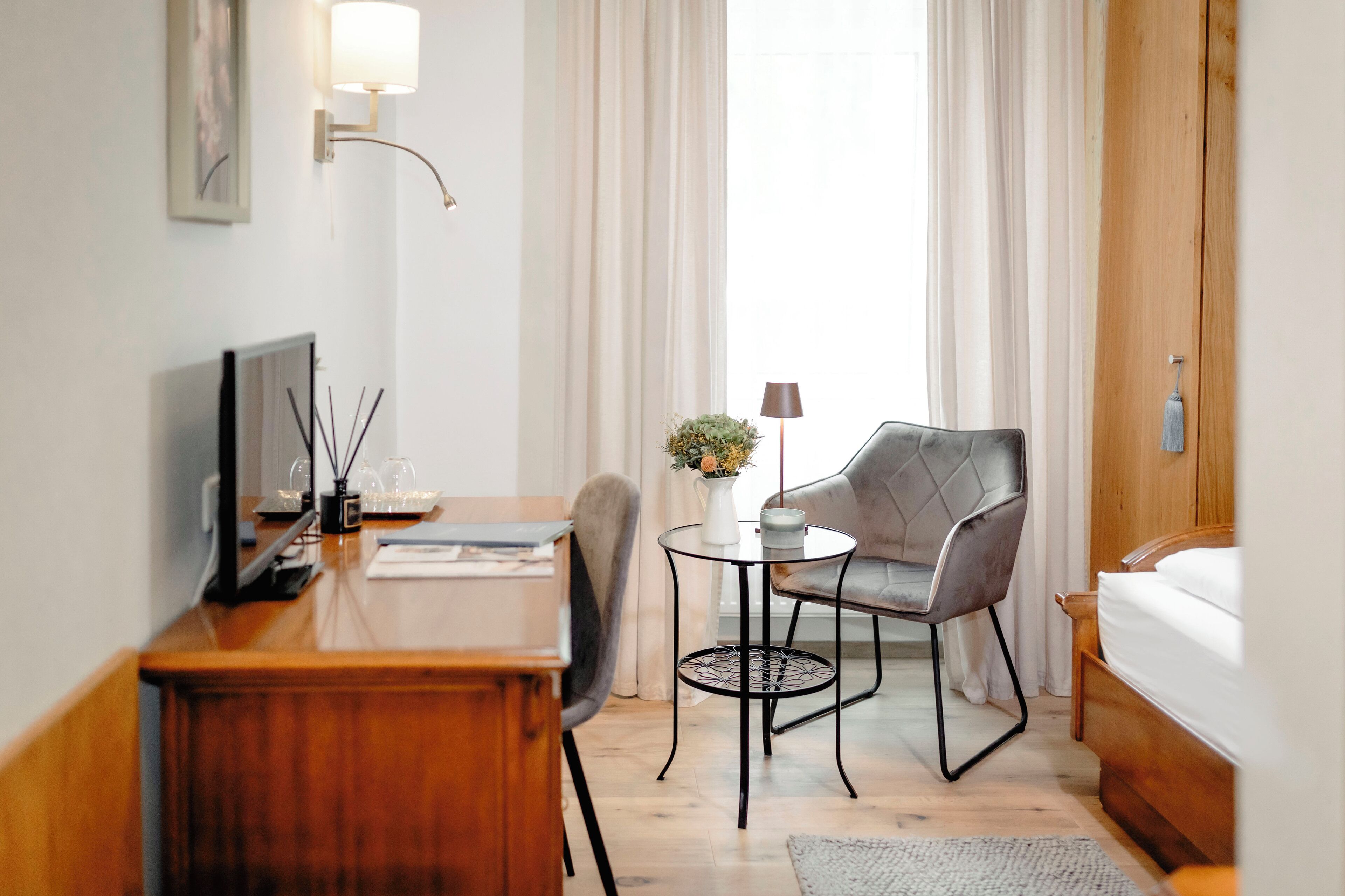 A hotel room with a wooden desk, a chair, a small round table with a vase, a lamp, and a bed.