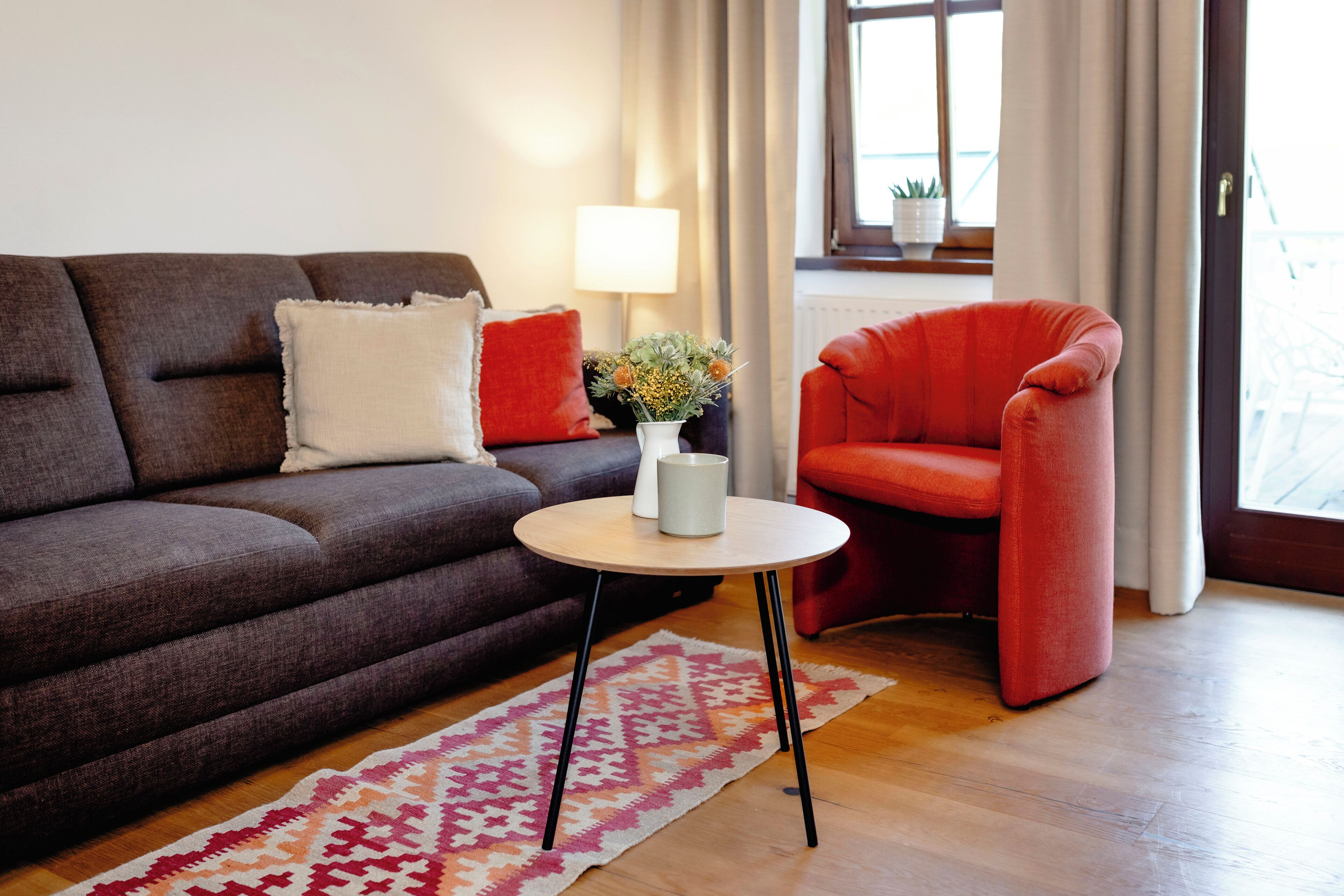 A living room with a brown sofa, a red armchair, a small round table with a vase, and a patterned rug.
