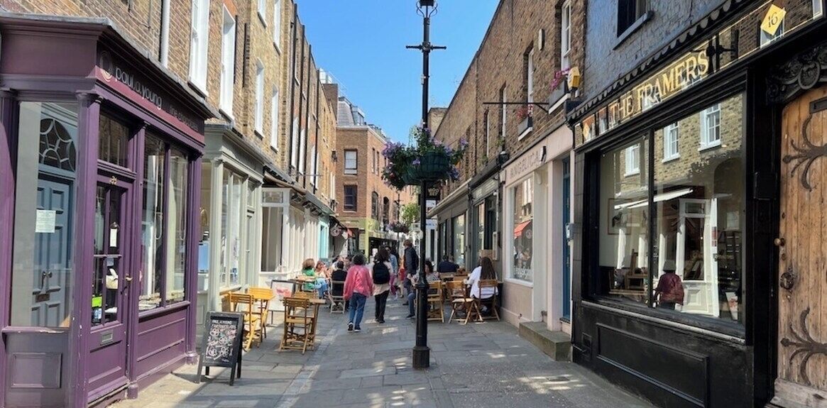 A narrow street lined with shops, including a visible cafe with outdoor seating.