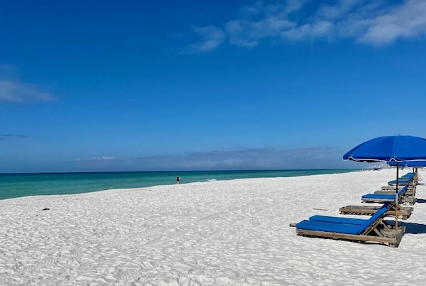 A beach with white sand, a row of blue umbrellas, and a person walking in the distance.
