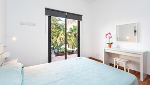 Une chambre à coucher comprenant un lit, un bureau, une chaise, une plante en pot et une porte coulissante donnant sur un balcon agrémenté de plantes et offrant une vue sur l’extérieur.