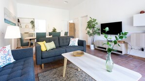 Un salon moderne avec un canapé gris, une table basse et un téléviseur. On aperçoit des plantes et une bibliothèque à l’arrière-plan.
