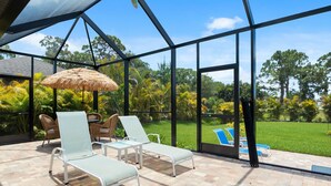 A covered patio with lounge chairs and a thatched umbrella, overlooking a lush garden and a swimming pool. - Coral Breeze  | Sunshelf | 4 Bed, 3 Bath (Rotonda West)