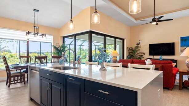 A modern kitchen with a large island, a dining area with chairs, a red sofa, and a TV mounted on the wall. - Coral Breeze  | Sunshelf | 4 Bed, 3 Bath (Rotonda West)