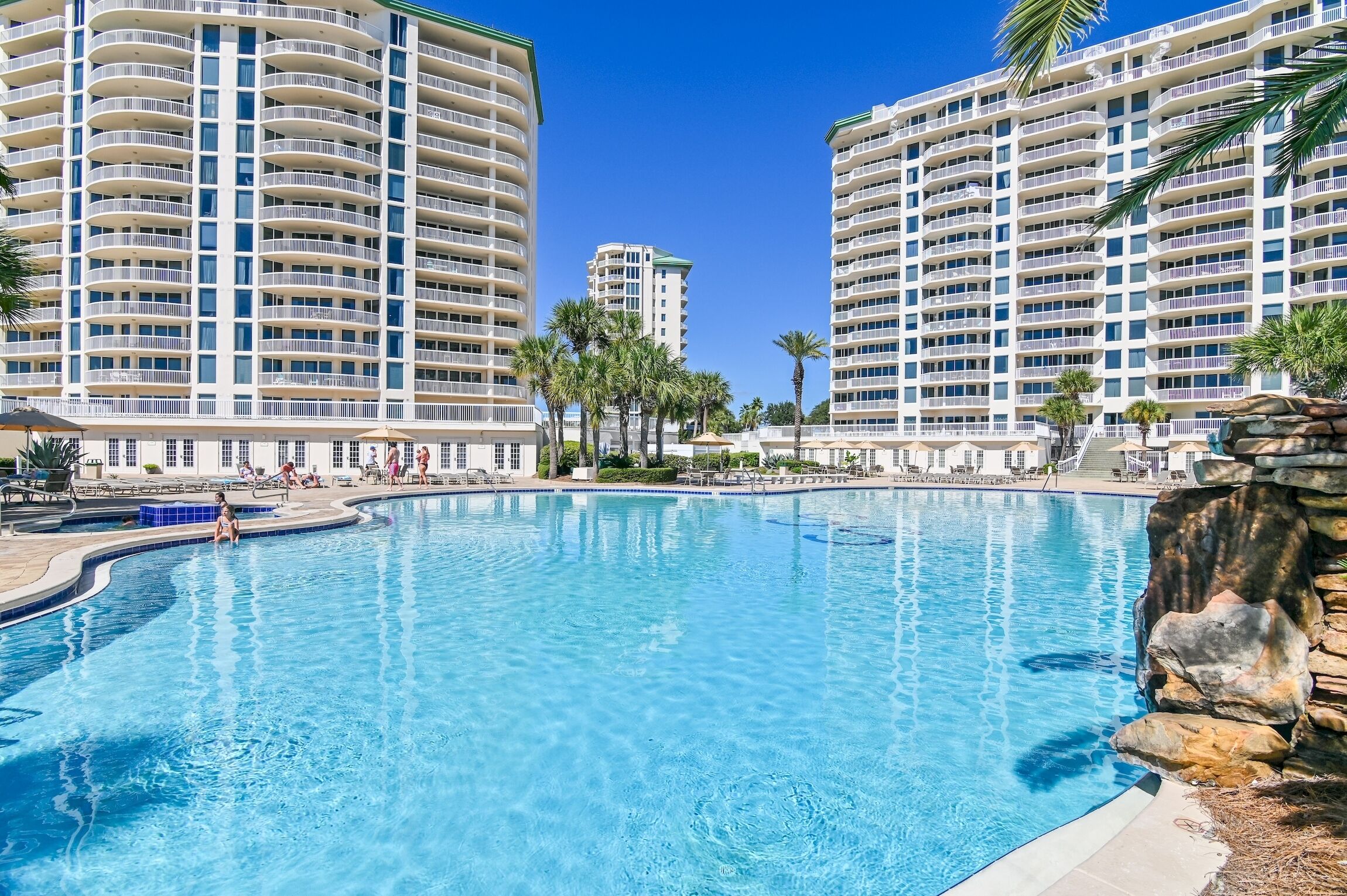 A large swimming pool surrounded by high-rise buildings with balconies, palm trees, and a clear blue sky.