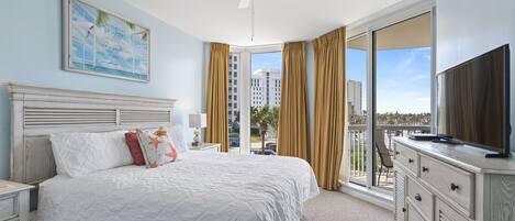 A hotel room with a large bed, a ceiling fan, a television on a dresser, and a large window with curtains.