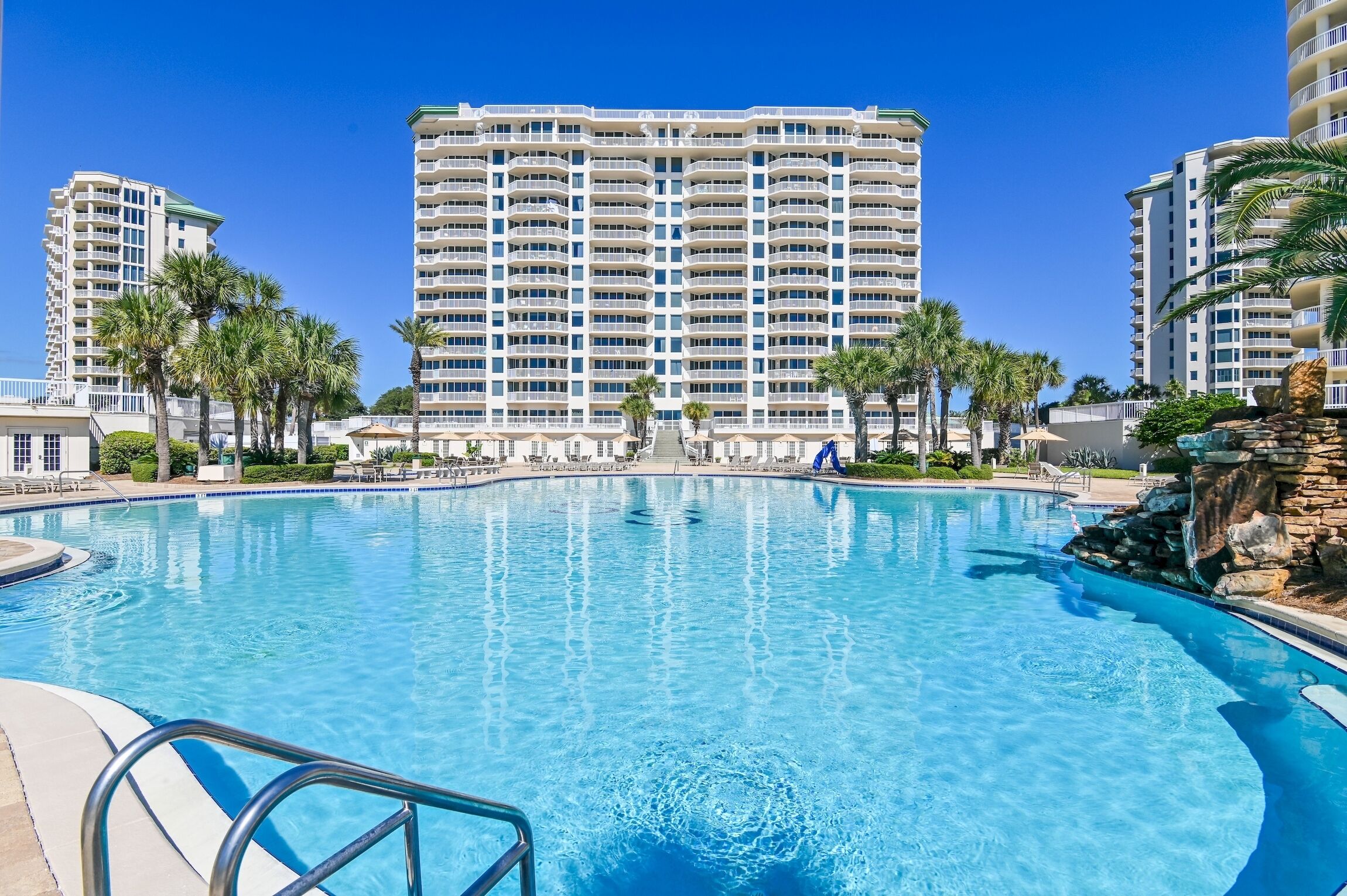 A large swimming pool with a clear blue water, surrounded by palm trees and a tall hotel building in the background.