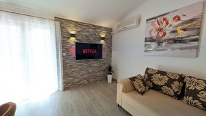 A living room with a stone wall, a flat-screen TV, a sofa with floral cushions, and a painting on the wall. - Easy City Access: 3BR w/ Balcony & Private Carport (Zadar)