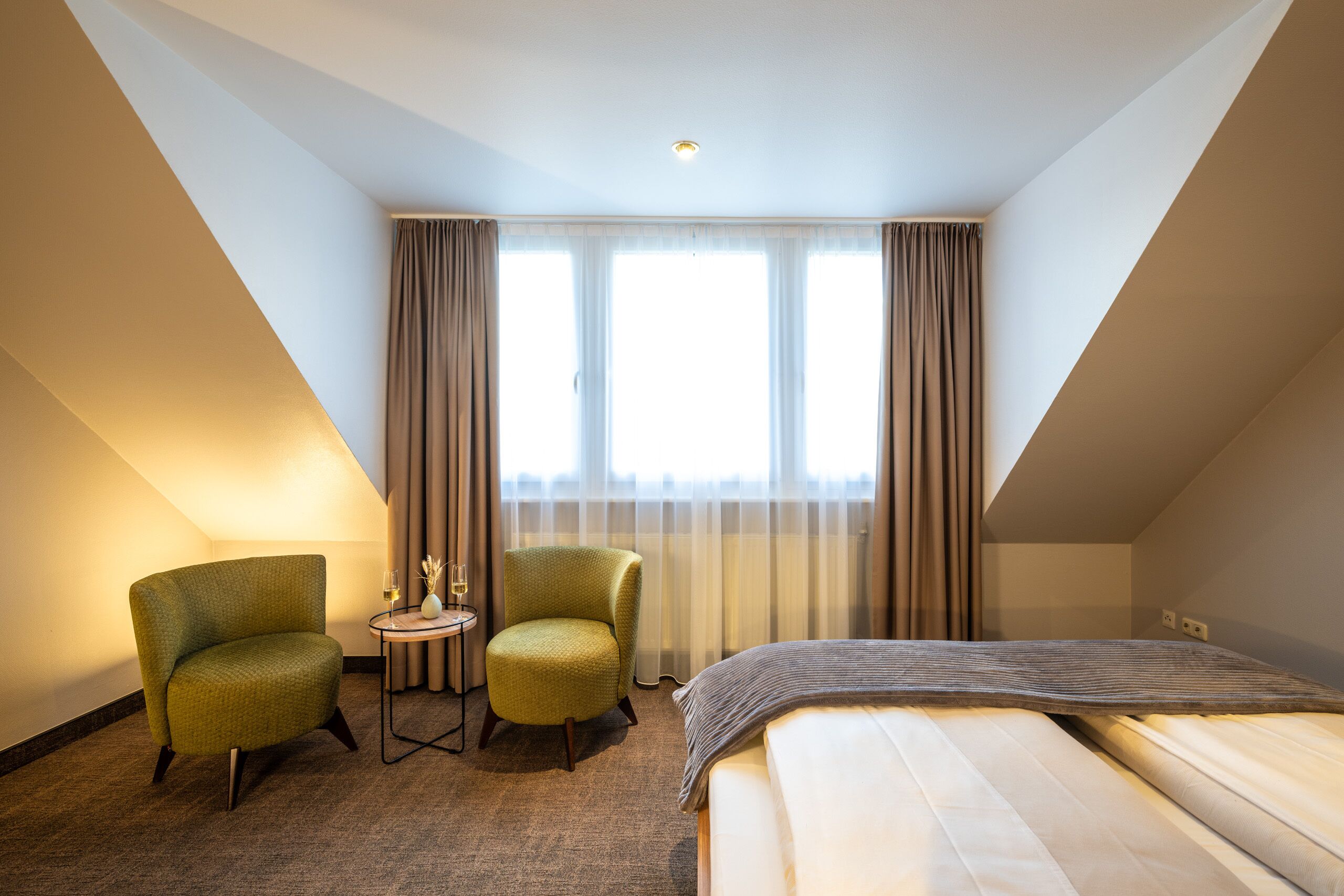 Een hotelkamer met een bed, twee fauteuils, een tafeltje met een glas wijn en een raam met gordijnen.