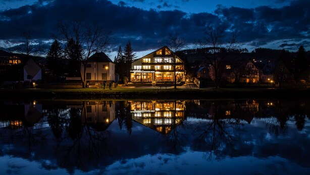 A large, illuminated building with a reflection in the water, surrounded by trees and other buildings. - Uferdeck - Boutique Hotel (Traben-Trarbach)