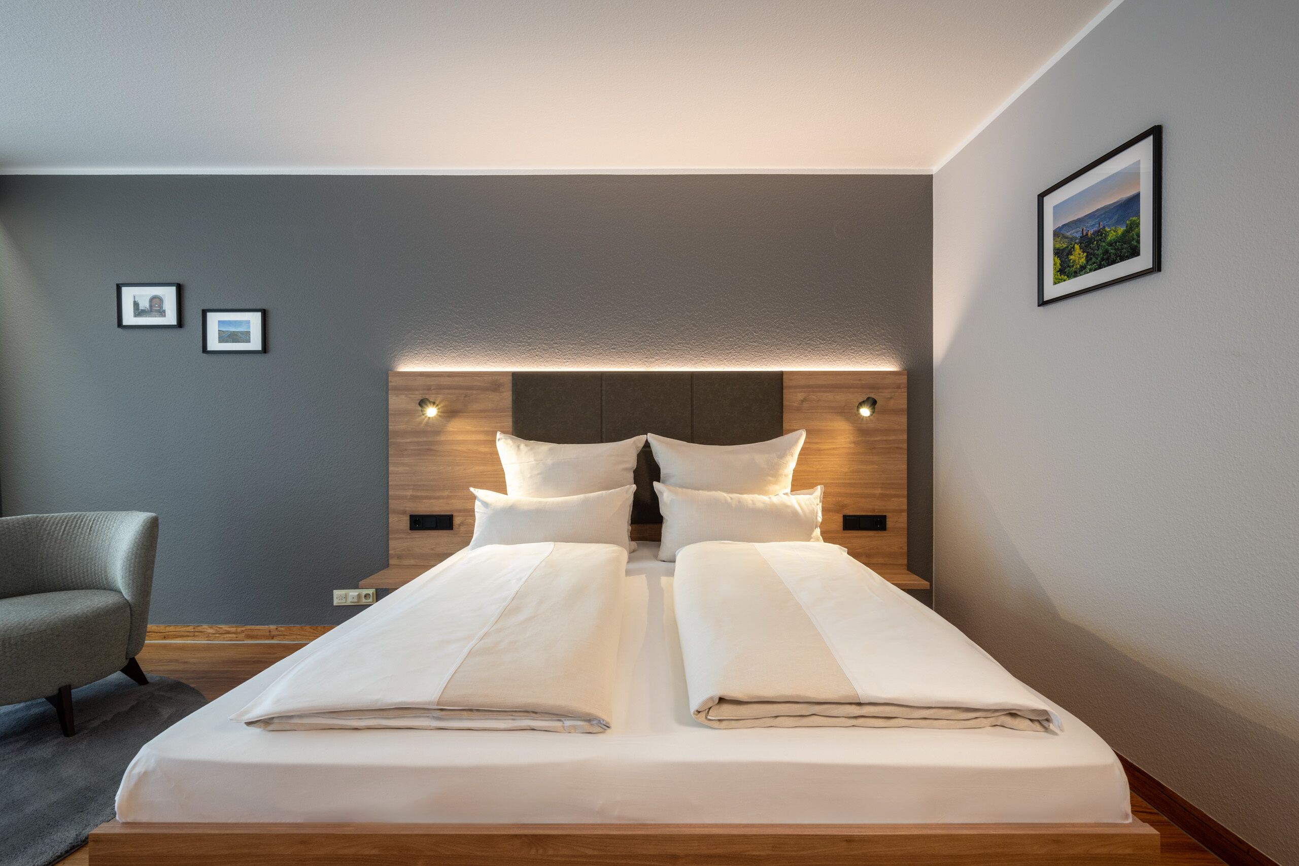 Una camera d'albergo moderna con un letto grande, una testiera in legno e un muro grigio con due quadri incorniciati.