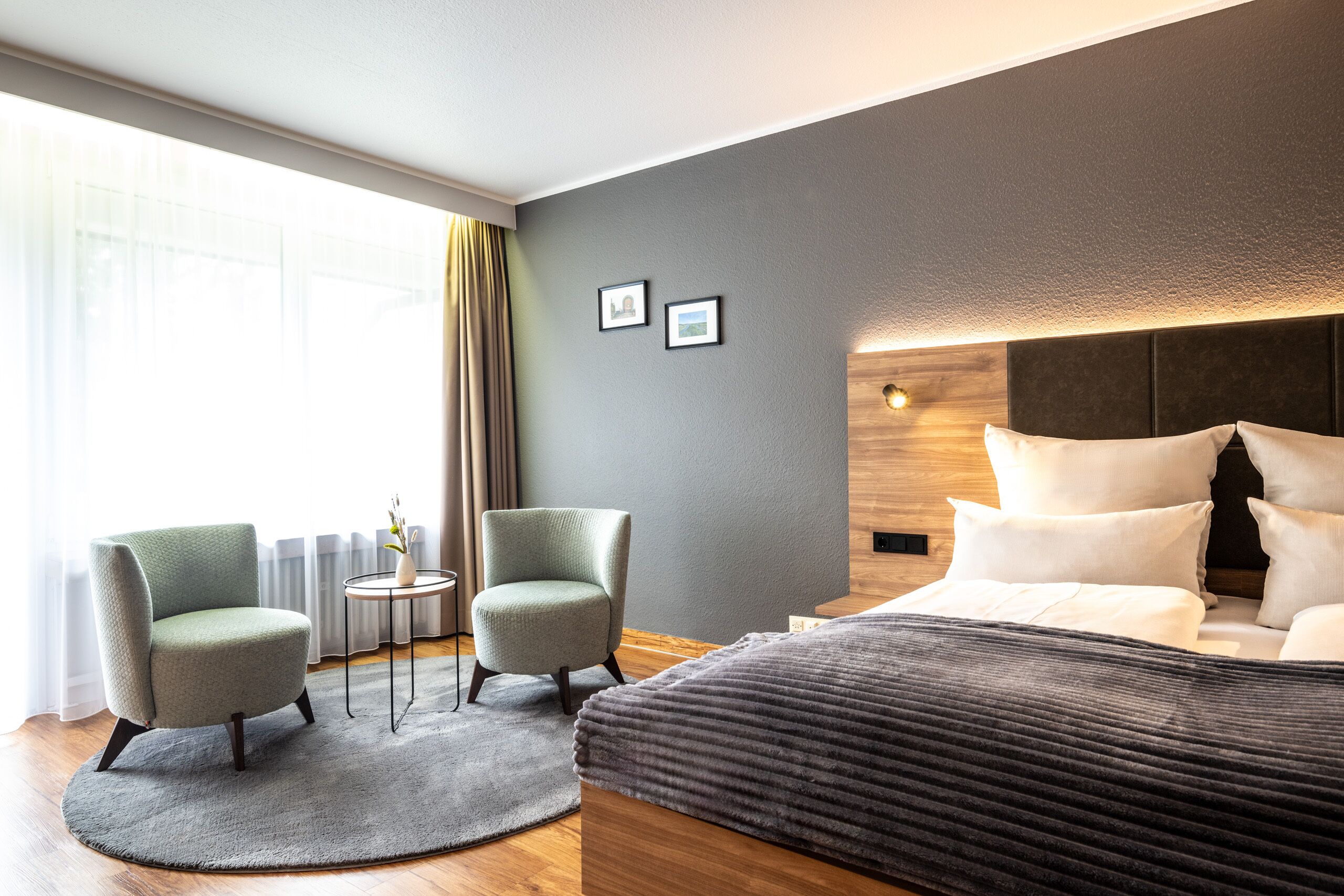 A modern hotel room with a large bed, two armchairs, a small side table, and a view of the city through large windows.