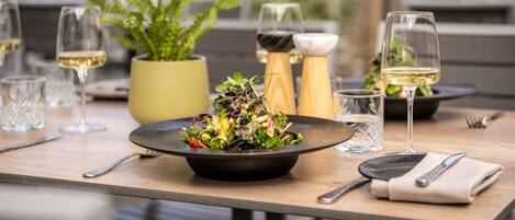 Una mesa de comedor con un bol negro de ensalada, copas de vino, cubiertos y una planta en maceta.