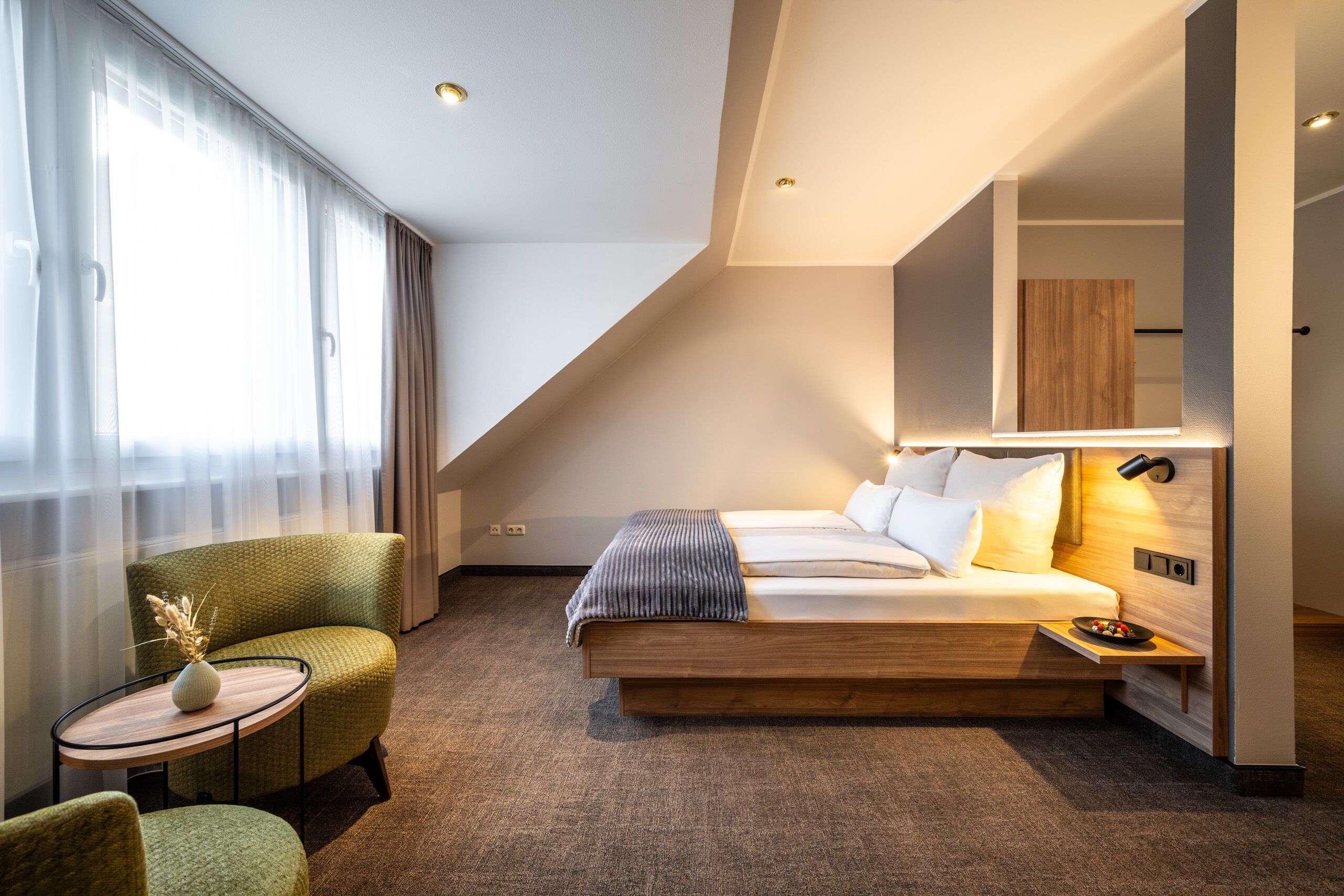 A modern hotel room with a bed, a small table with a vase, a chair, and a wardrobe.