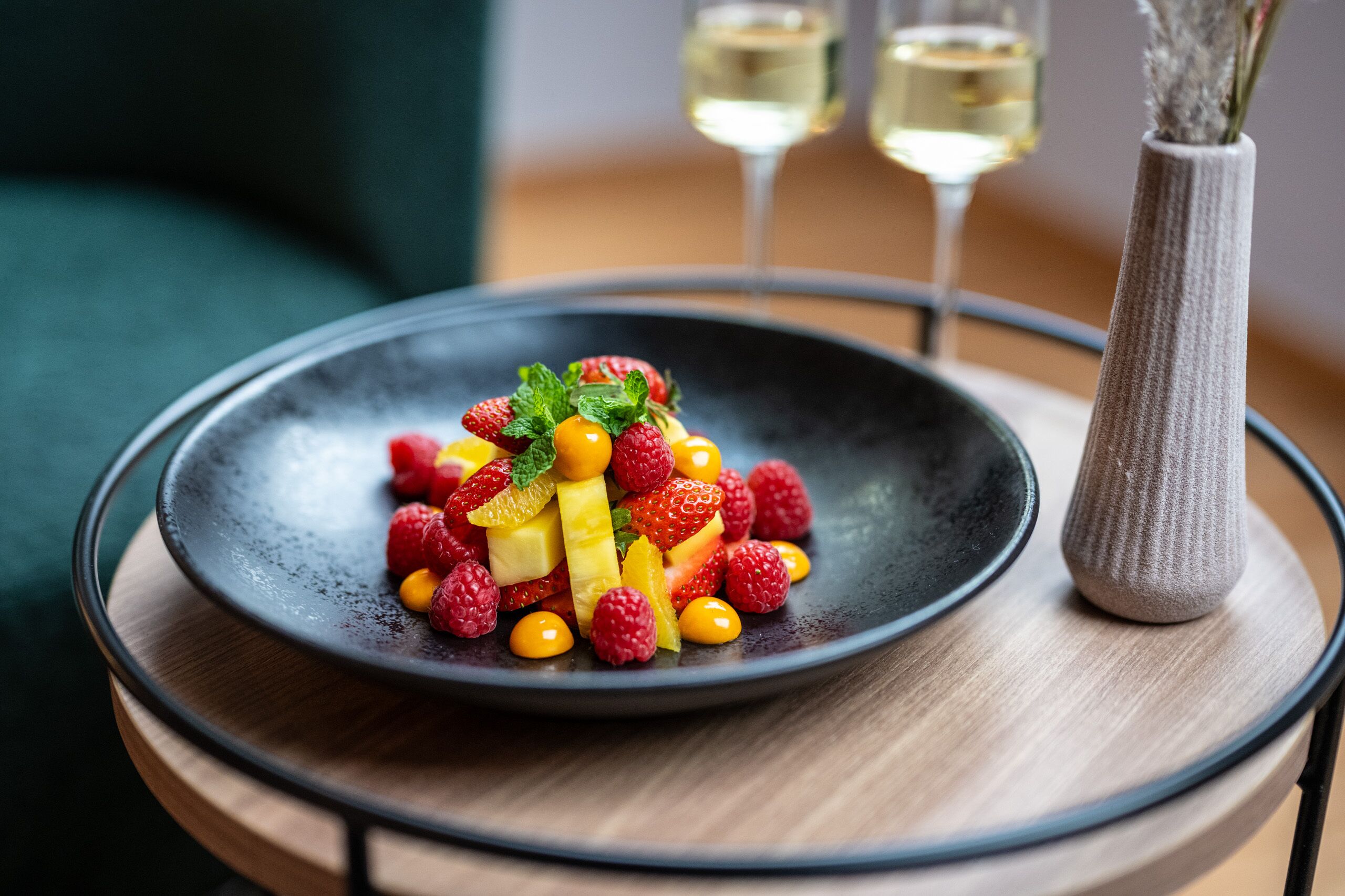 A fruit salad on a black plate with a vase and two glasses of white wine in the background.