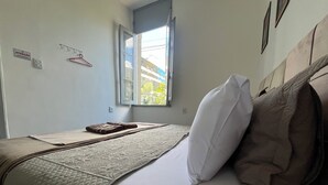 A neatly made bed with a quilt, pillows, and a folded towel. There is a window with a view of buildings outside. - Rota BH Hostel (Belo Horizonte)